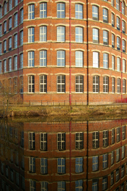 Curved red-brick end and repeated windows of Anchor Mill in Paisley, reflected in the water below